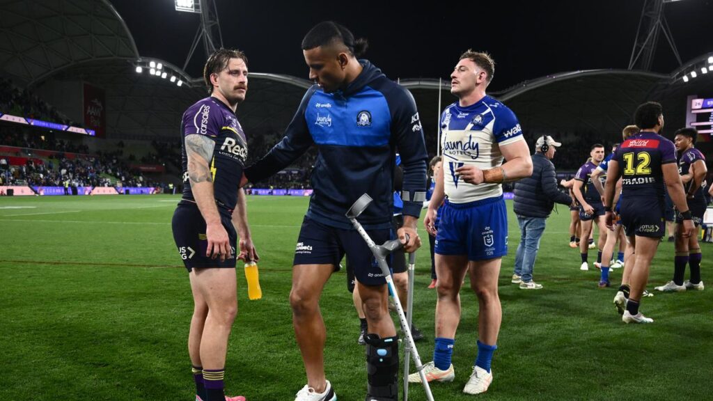 Bulldogs’ Stephen Crichton leaves AAMI Park on crutches to sympathy from Storm’s Cameron Munster.
